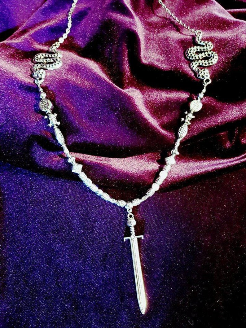May include: A silver necklace with a sword pendant and two snake charms. The necklace is on a purple velvet background.