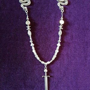 May include: A silver necklace with two snake charms, a sword pendant, and silver beads. The necklace is on a purple background.