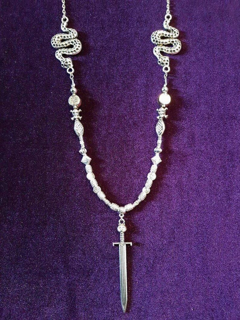 May include: A silver necklace with two snake charms, a sword pendant, and silver beads. The necklace is on a purple background.