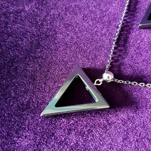 May include: A silver chain necklace with a silver triangle pendant. The pendant has a hollow center and is hanging on a purple velvet surface.