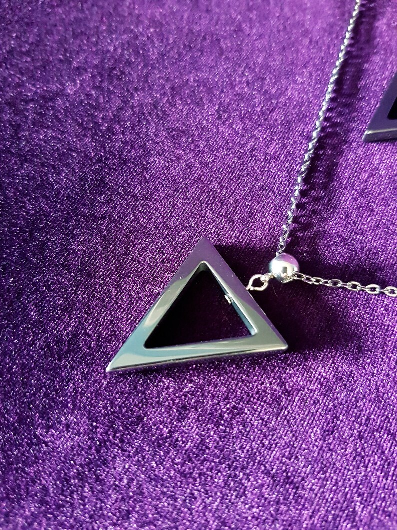 May include: A silver chain necklace with a silver triangle pendant. The pendant has a hollow center and is hanging on a purple velvet surface.