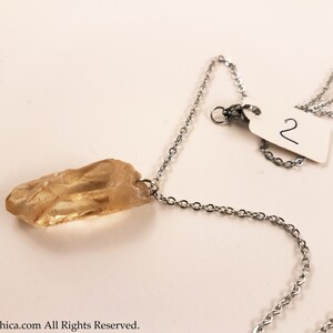 May include: A silver chain necklace with a light brown crystal pendant. The pendant is a long, thin, and pointed crystal. The necklace has a tag with the number "2" on it.