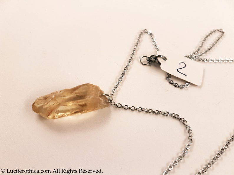May include: A silver chain necklace with a light brown crystal pendant. The pendant is a long, thin, and pointed crystal. The necklace has a tag with the number "2" on it.
