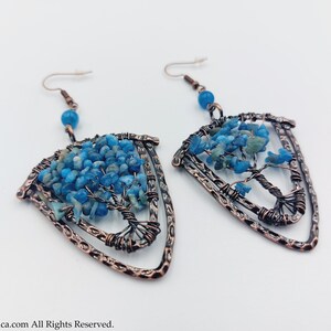 May include: A pair of copper wire earrings featuring a tree of life design. The tree is crafted from blue and green stones and is enclosed within a copper wire frame. The earrings are shaped like a shield.