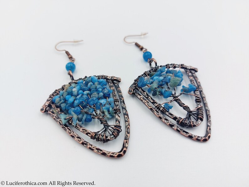 May include: A pair of copper wire earrings featuring a tree of life design. The tree is crafted from blue and green stones and is enclosed within a copper wire frame. The earrings are shaped like a shield.