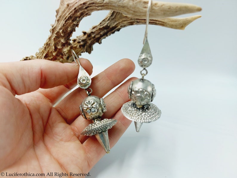 May include: A pair of silver dangle earrings with a round, textured ball and a pointed, textured cone. The earrings are hanging from a silver hook.