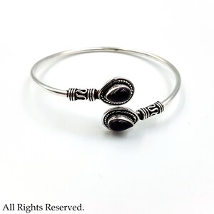 May include: A silver open cuff bracelet featuring two teardrop-shaped dark purple stones at the ends. The metal band displays intricate etched detailing near the settings. The text Luciferthica.com All Rights Reserved is at the bottom.