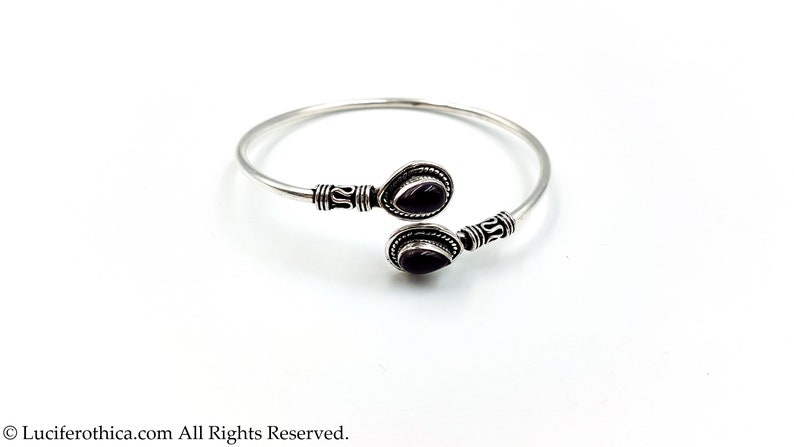 May include: A silver open cuff bracelet featuring two teardrop-shaped dark purple stones at the ends. The metal band displays intricate etched detailing near the settings. The text Luciferthica.com All Rights Reserved is at the bottom.