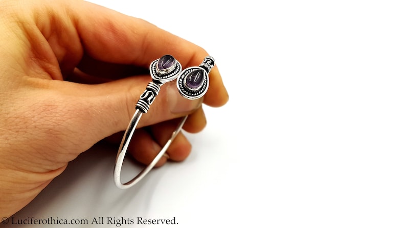 May include: A silver-toned open cuff bracelet featuring two teardrop-shaped purple amethyst stones set in decorative metal bezels. The band has a smooth finish with textured, dark-oxidized metal accents near the stone settings at each end.
