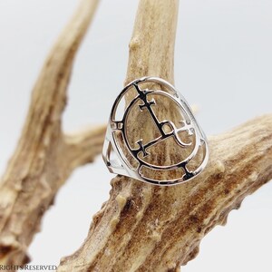 May include: A silver ring with a circular design featuring a cross and other symbols. The ring is displayed on a piece of wood.