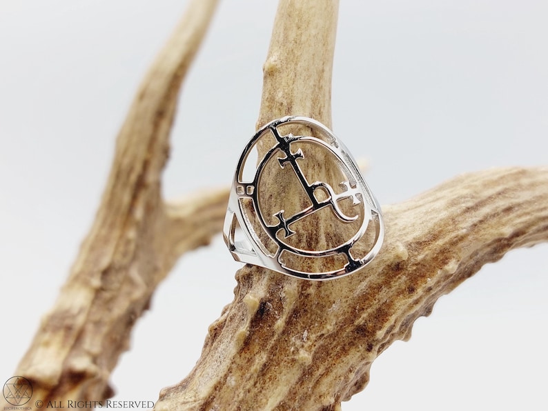 May include: A silver ring with a circular design featuring a cross and other symbols. The ring is displayed on a piece of wood.