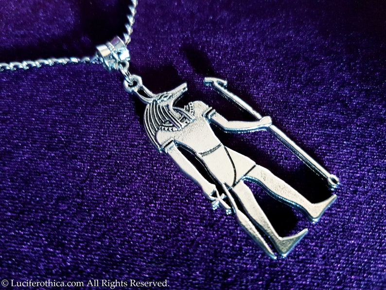 May include: Silver pendant necklace featuring an Egyptian god Anubis with a jackal head, holding a staff. The pendant is on a purple velvet background.
