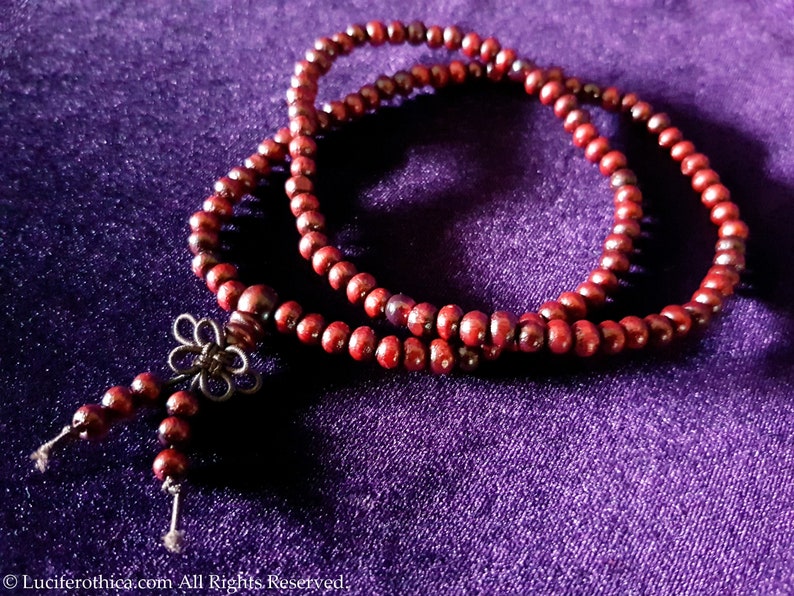 Op de afbeelding: Een donkerrode houten mala gebedskralenketting met een zilveren bloemenbedel. De ketting ligt op een paarse fluwelen ondergrond.