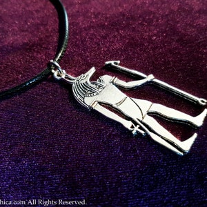 May include: Silver pendant necklace featuring an Anubis figure holding a staff. The pendant is on a black leather cord.