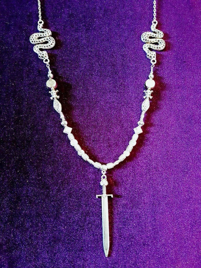 May include: A silver necklace with two snake charms and a sword pendant. The necklace is on a purple background.