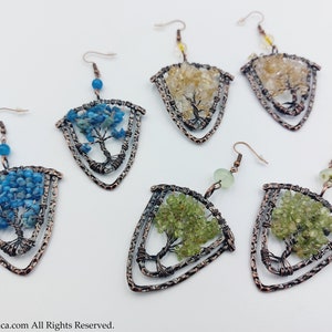 May include: Six pairs of copper wire tree of life earrings with different coloured gemstones. The earrings are in the shape of a triangle with a tree design in the centre. The gemstones are blue, green, and yellow.