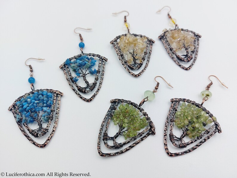 May include: Six pairs of copper wire tree of life earrings with different coloured gemstones. The earrings are in the shape of a triangle with a tree design in the centre. The gemstones are blue, green, and yellow.