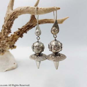 May include: A pair of silver dangle earrings with a teardrop-shaped top, a round bead with floral designs, and a pointed cone-shaped bottom.