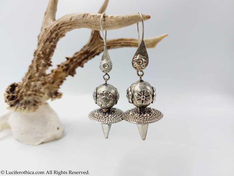 May include: A pair of silver dangle earrings with a teardrop-shaped top, a round bead with floral designs, and a pointed cone-shaped bottom.