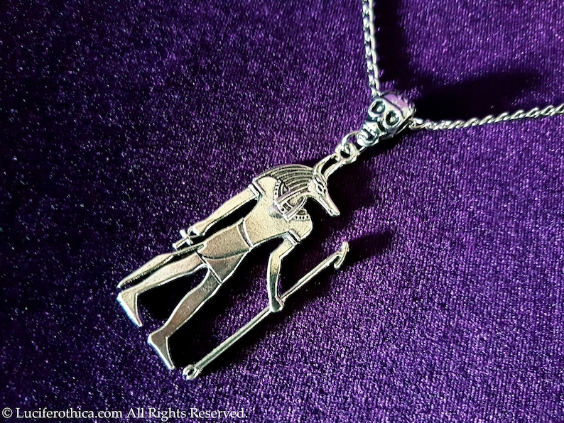 May include: Silver necklace featuring a pendant depicting an Egyptian god with a jackal head. The pendant is detailed and intricate, with the god holding a staff in one hand.