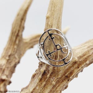 May include: A silver ring with a circular design featuring a symbol with crosses and a crescent moon. The ring is displayed on a piece of antler.