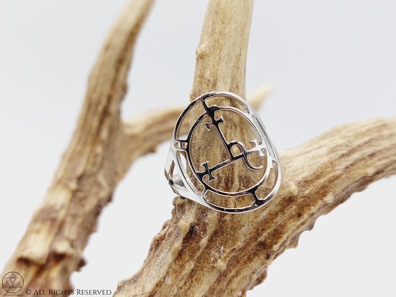 May include: A silver ring with a circular design featuring a symbol with crosses and a crescent moon. The ring is displayed on a piece of antler.