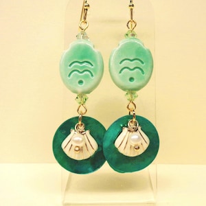 Può includere: Un paio di orecchini pendenti con un pezzo in ceramica verde menta chiaro con motivo a onde, collegato da un piccolo cristallo verde a un disco circolare verde ottanio scuro. Un ciondolo a forma di conchiglia bianca con una piccola perla.