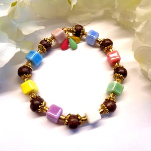 Peut inclure: Un bracelet coloré avec des perles carrées de différentes couleurs, dont le bleu, le rose, le jaune et le vert, alternant avec des perles marron foncé et des détails dorés. Le bracelet comporte de petits charms pendants et est posé sur un fond blanc.