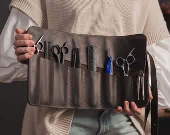 Puede incluir: Un estuche enrollable de cuero marrón con múltiples bolsillos para guardar herramientas de peluquería. El estuche está abierto y contiene una variedad de tijeras, peines y navajas.