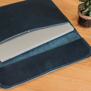 May include: A dark blue leather laptop sleeve with a silver laptop inside. The sleeve has white stitching around the edges and is open at the top. The laptop is partially visible, and the sleeve is on a wooden surface.