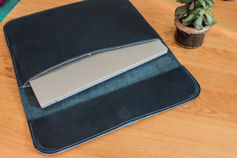 May include: A dark blue leather laptop sleeve with a silver laptop inside. The sleeve has white stitching around the edges and is open at the top. The laptop is partially visible, and the sleeve is on a wooden surface.