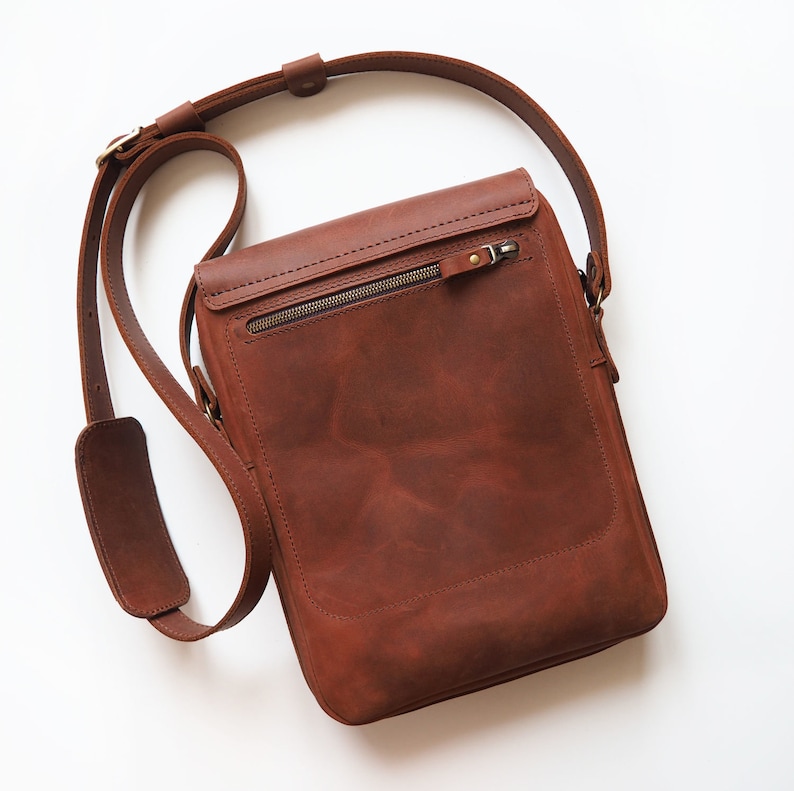 Puede incluir: Un bolso de mensajero de cuero marr&oacute;n con un bolsillo con cremallera en la parte delantera. El bolso tiene una correa larga ajustable.