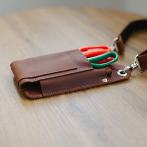 May include: A brown leather tool belt with a strap and three pairs of scissors in a pocket. The scissors are red, green, and green.
