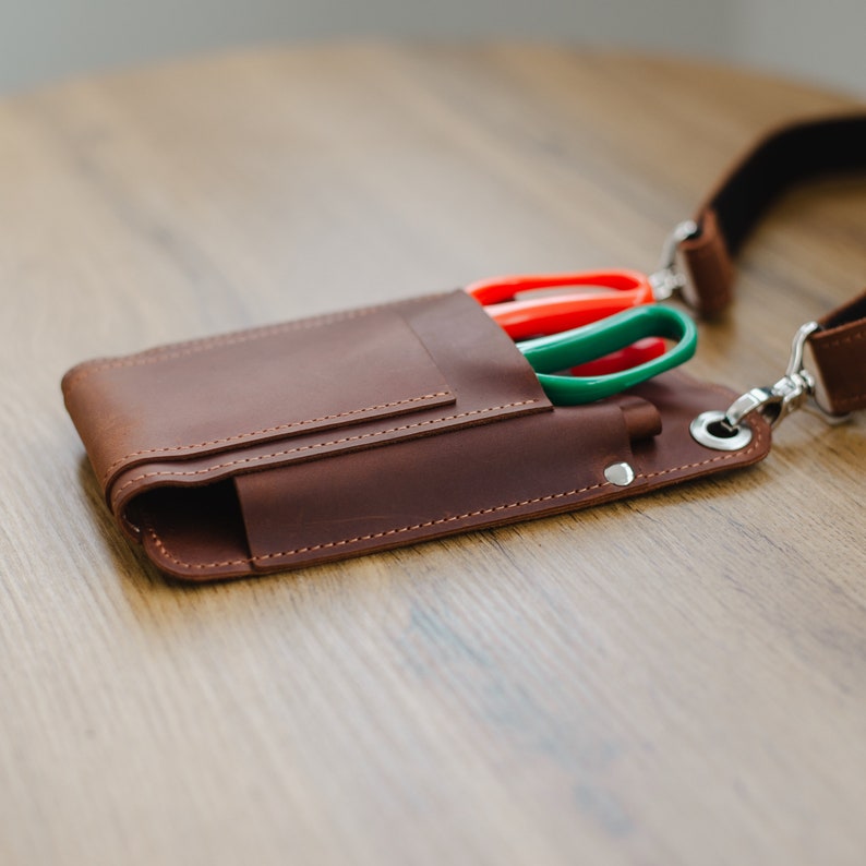 May include: A brown leather tool belt with a strap and three pairs of scissors in a pocket. The scissors are red, green, and green.