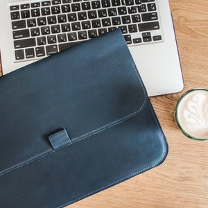 May include: A navy blue leather laptop sleeve with a clasp closure rests on a wooden desk next to a silver laptop and a glass of latte art. The sleeve is rectangular and has a stitched border.