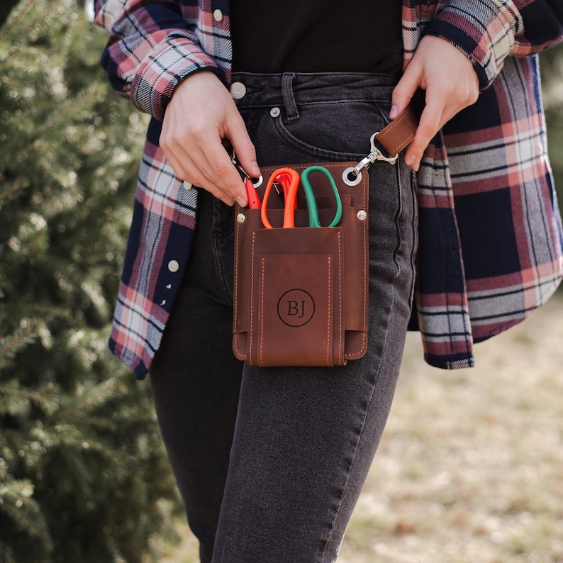 May include: A brown leather tool belt with a pocket for carrying gardening shears. The belt has a metal buckle and a personalized monogram "BJ".