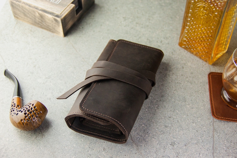May include: A brown leather tobacco pouch with a strap closure. The pouch is rolled up and tied with the strap.