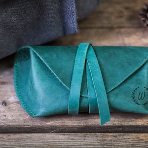 K&ouml;nnte beinhalten: Tealfarbene Leder-Brillentasche mit gr&uuml;ner Naht und einem Monogramm "W" in einem Kranz-Design.
