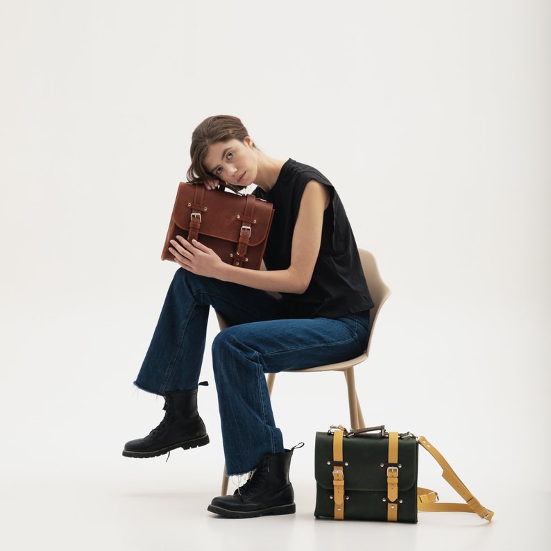 K&ouml;nnte beinhalten: Eine Frau sitzt auf einem Stuhl und h&auml;lt eine braune Ledertasche. Eine zweite gr&uuml;ne Ledertasche mit gelben Akzenten steht vor ihr auf dem Boden.