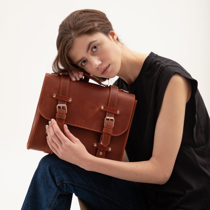 K&ouml;nnte beinhalten: Eine braune Ledertasche mit mehreren Schnallen und Riemen. Die Tasche wird von einer Person gehalten, die ein schwarzes &auml;rmelloses Hemd und eine blaue Jeans tr&auml;gt.