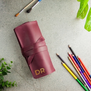 May include: A burgundy leather roll-up case for pencils or brushes, featuring a wrap-around leather tie closure and the gold-coloured letters DR embossed on the front surface. The case is positioned alongside several coloured pencils and paintbrushes.