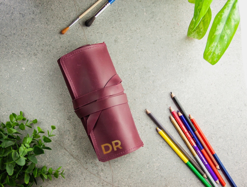 May include: A burgundy leather roll-up case for pencils or brushes, featuring a wrap-around leather tie closure and the gold-coloured letters DR embossed on the front surface. The case is positioned alongside several coloured pencils and paintbrushes.