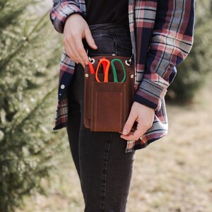 May include: A brown leather tool belt with a flap closure and three pockets. The belt is worn over a pair of dark denim jeans and a plaid shirt. The pockets contain a pair of orange and green pruning shears.