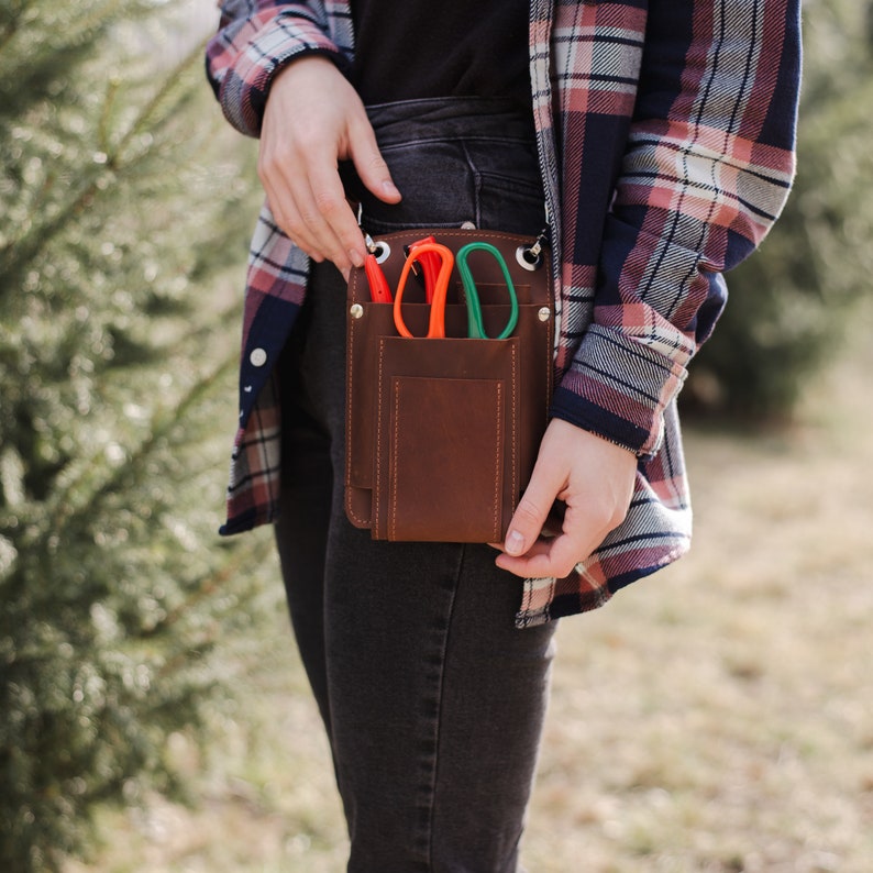 May include: A brown leather tool belt with a flap closure and three pockets. The belt is worn over a pair of dark denim jeans and a plaid shirt. The pockets contain a pair of orange and green pruning shears.