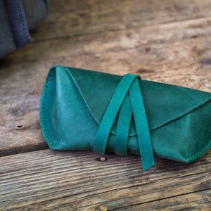K&ouml;nnte beinhalten: Ein Brillenetui aus tealgr&uuml;nem Leder mit Klappenverschluss und Bindeverschluss. Das Etui ist aus weichem Leder gefertigt und hat eine glatte Oberfl&auml;che.