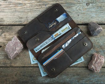 Puede incluir: Cartera de cuero marrón abierta con múltiples compartimentos, incluyendo ranuras para tarjetas y un bolsillo con cremallera. La cartera contiene efectivo y tarjetas de crédito. La cartera se muestra abierta sobre una superficie de madera, con rocas decorativas.