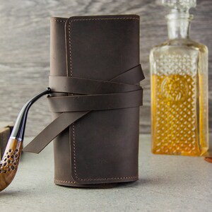 May include: A brown leather pipe roll with a strap closure. The roll is closed and sitting on a table next to a glass of whiskey and a decanter. A wooden pipe is lying on the table in front of the roll.