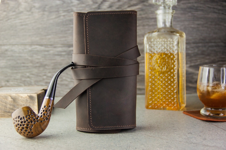 May include: A brown leather pipe roll with a strap closure. The roll is closed and sitting on a table next to a glass of whiskey and a decanter. A wooden pipe is lying on the table in front of the roll.