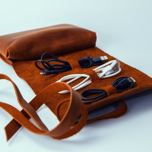May include: A brown leather roll-up case for organizing and storing cords and cables. The case has multiple slots for holding different types of cords and cables.