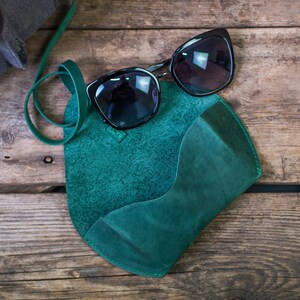 K&ouml;nnte beinhalten: Ein Sonnenbrillenetui aus tealgr&uuml;nem Leder mit einem Riemen. Das Etui liegt auf einer Holzoberfl&auml;che mit einer schwarzen Sonnenbrille darauf.